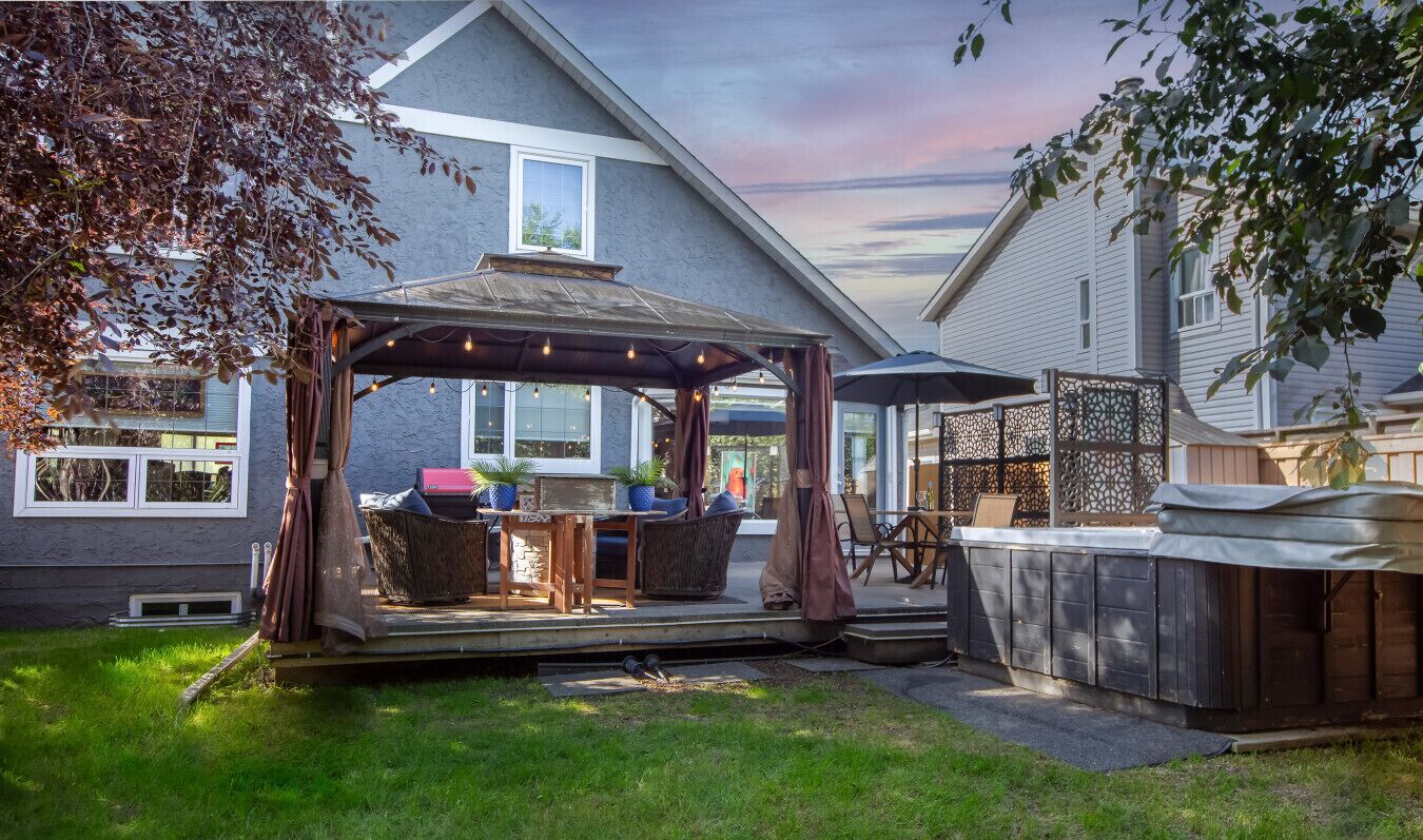 Un patio techado con mobiliario exterior, un jacuzzi y vista a una casa al fondo.
