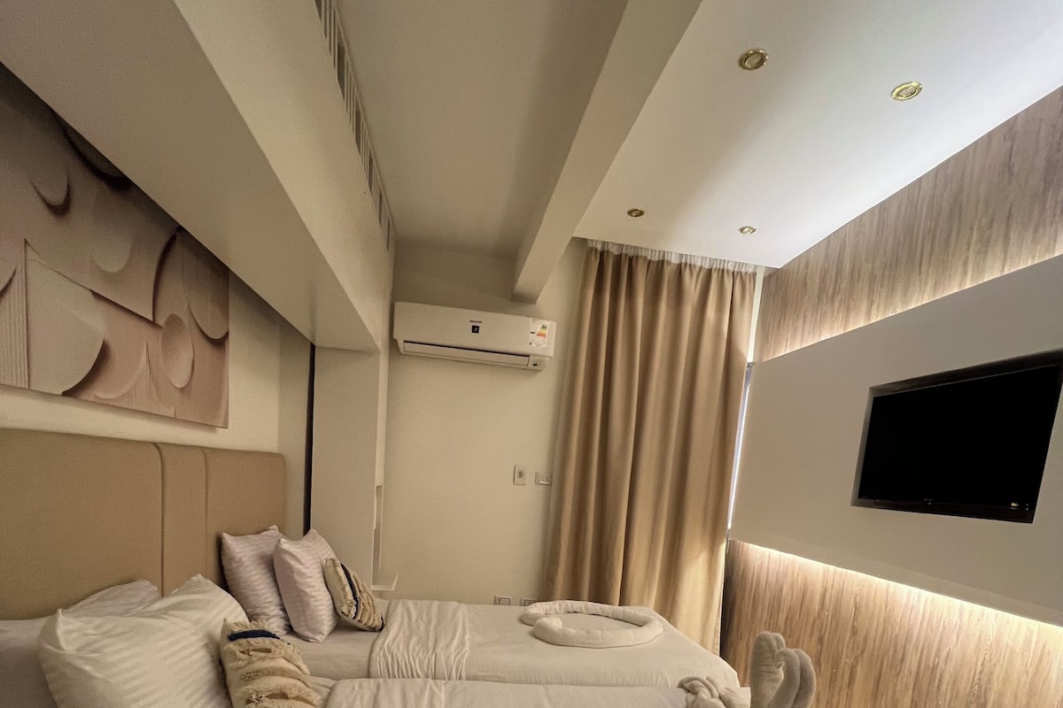 A modern hotel room with a large bed, a flat-screen TV mounted on the wall, and a ceiling air conditioning unit.