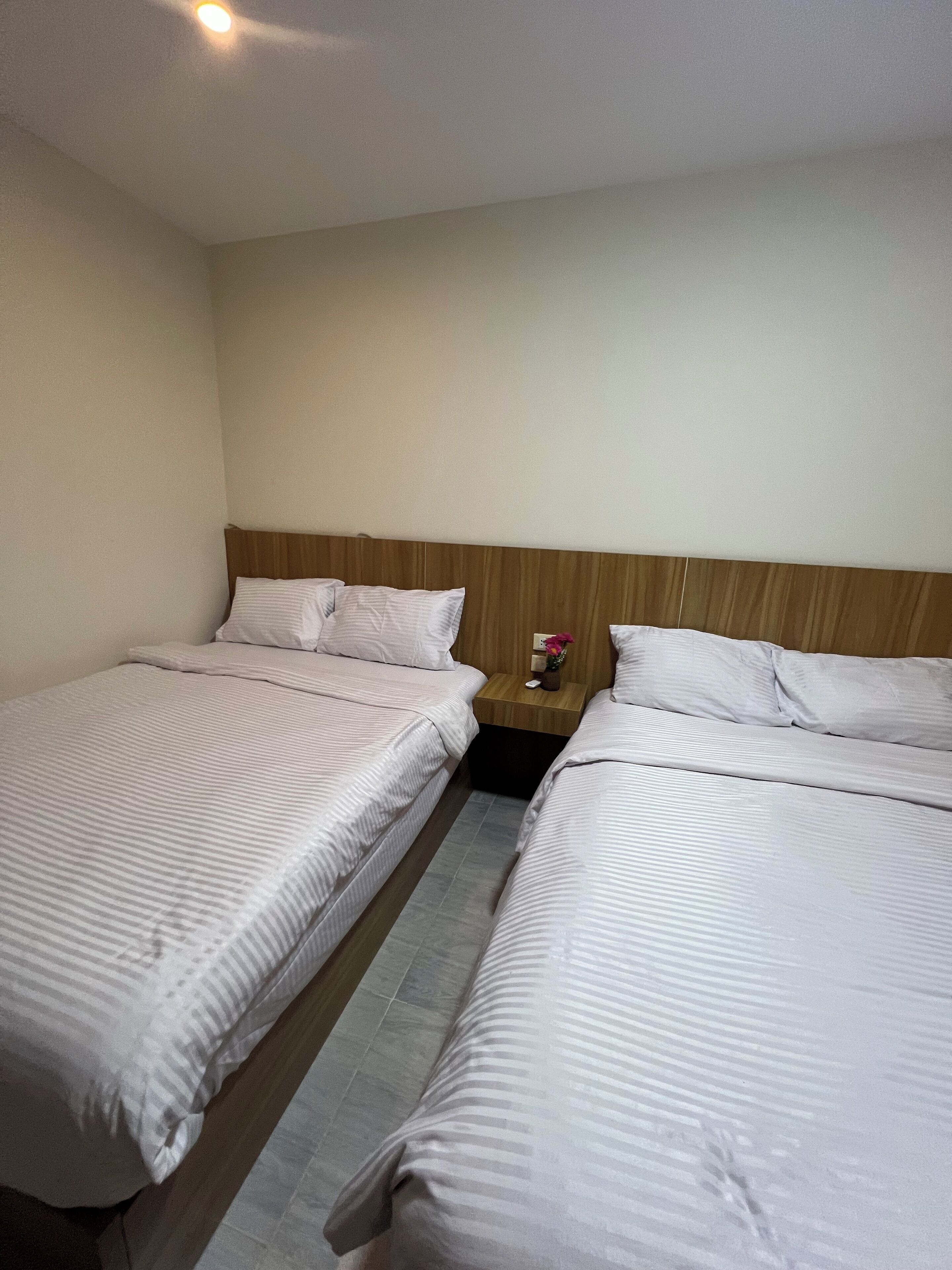 Habitación de hotel con dos camas individuales, cabecera de madera, mesita de noche con un florero y suelo de baldosas.