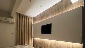 Moderni hotellihuone, jossa on suuri sänky, seinälle asennettu taulutelevisio ja ilmastointilaite sängyn yläpuolella.