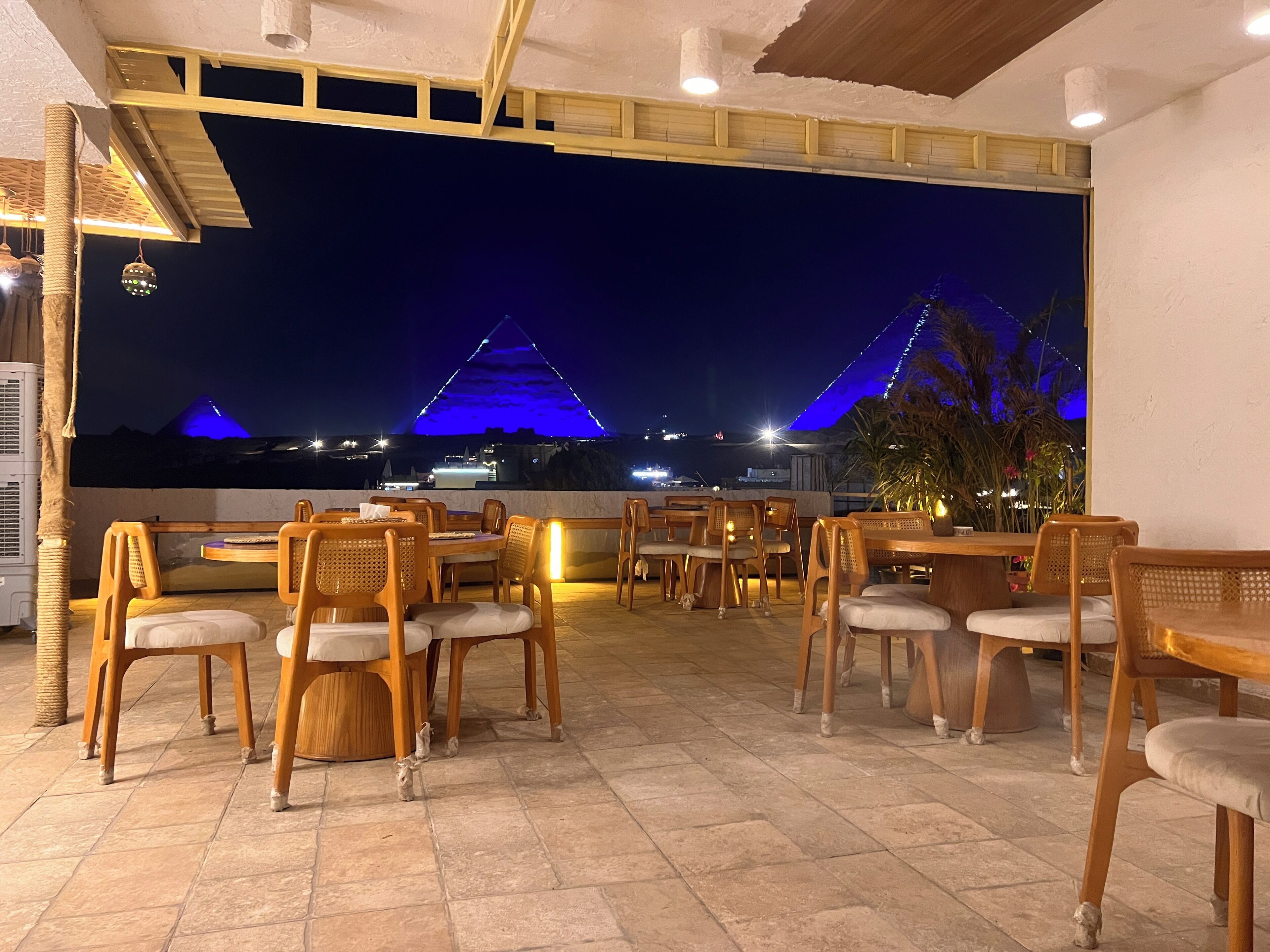An outdoor dining area with wooden tables and chairs, a view of a lit-up pyramid structure at night, and a ceiling with exposed beams and lighting fixtures.