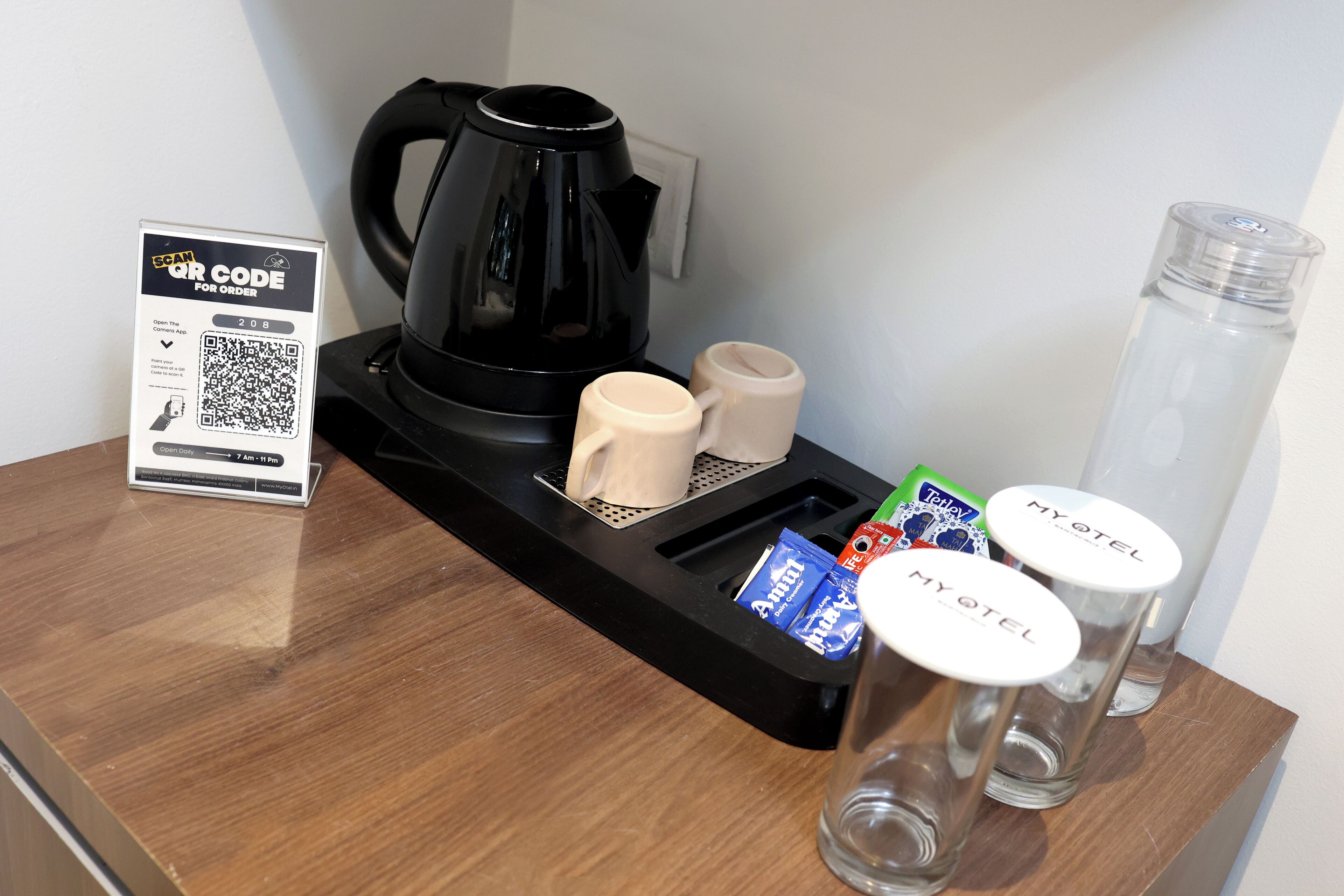 Montaje de desayuno en una habitación de hotel con una cafetera negra, dos recipientes para leche, una bandeja con sobres de café y dos vasos que dicen "MY OTEL".