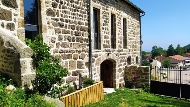 A stone building with a wooden fence, a small garden, and a paved area with a yellow car.