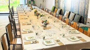 A long dining table set for a meal with chairs on both sides, a view of the outdoors through a large window, and a wall with a forest-themed artwork.
