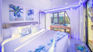 A bedroom with a bed, pillows, a nightstand, a window, and artwork on the wall. - Kokomo Floating Bungalow (Merritt Island)