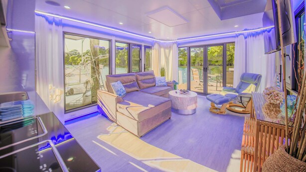A modern living room with a sofa, chairs, a coffee table, and large windows offering a view of the outdoors. - Kokomo Floating Bungalow (Merritt Island)