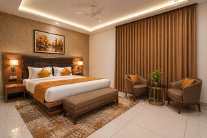 Una habitación de hotel moderna con una cama grande, dos mesitas de noche con lámparas, un ventilador de techo, un ventanal con cortinas y una pequeña zona de estar con dos sillones y una mesa.