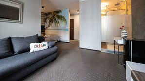 A modern hotel room with a grey sofa, a painting of a beach, a small bathroom with a shower, and a vase with flowers on a side table.