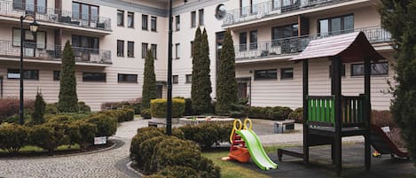 A courtyard with a playground, including a slide and a bench, surrounded by neatly trimmed bushes and a cobblestone path.