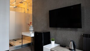 A modern bathroom with a glass shower enclosure, a black and white striped vase, a small potted plant, a black kettle, and a flat-screen TV mounted on the wall.