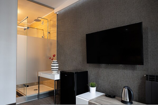 Un baño moderno con cabina de ducha de vidrio, un jarrón blanco y negro a rayas, una pequeña planta en maceta, una tetera negra y un televisor de pantalla plana montado en la pared.