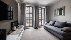 A modern living room with a grey sofa, a flat-screen TV mounted on the wall, a coffee table, and a large window with curtains.