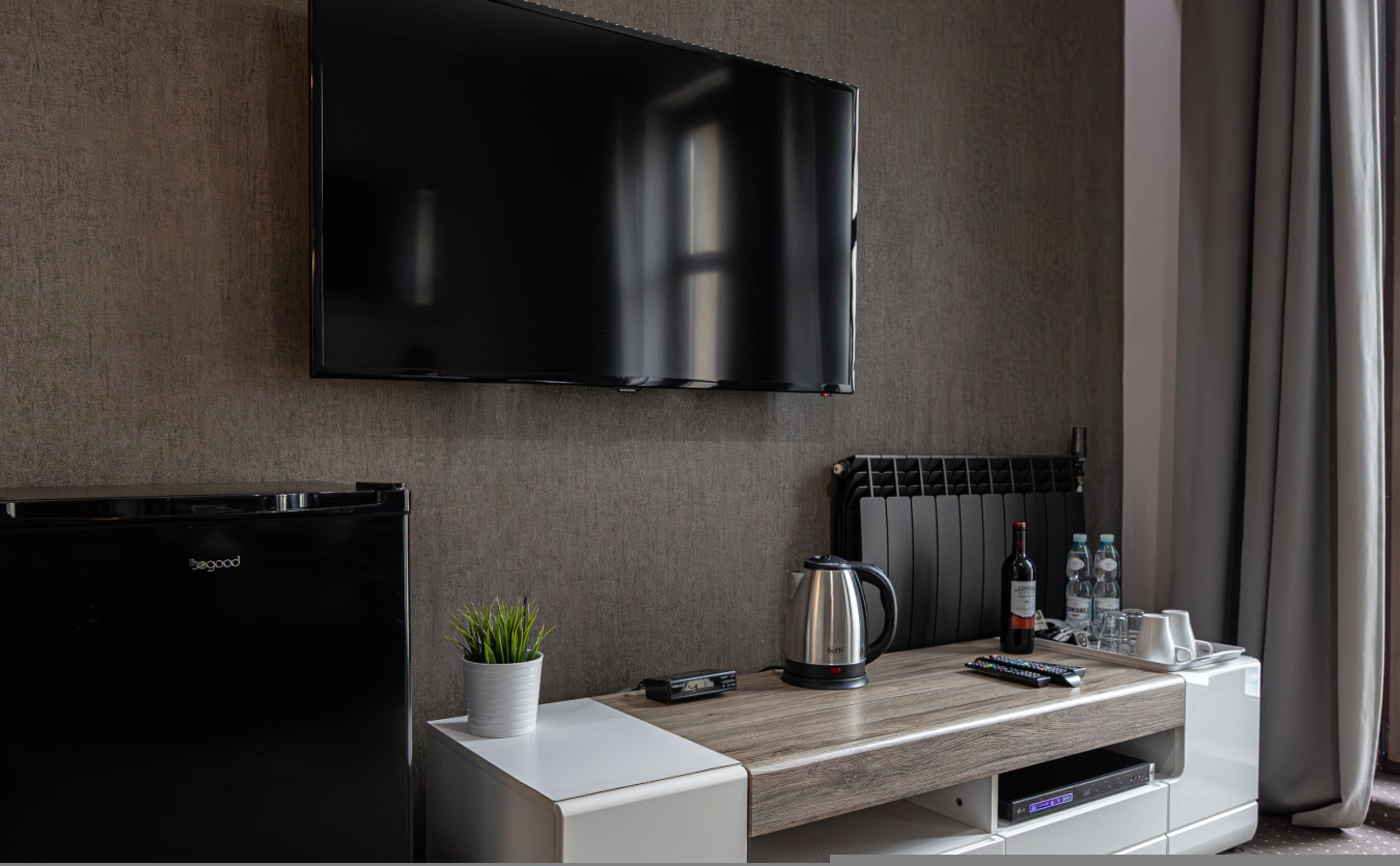 A modern hotel room with a flat-screen TV mounted on the wall, a minibar, a coffee maker, and a small potted plant on the counter.