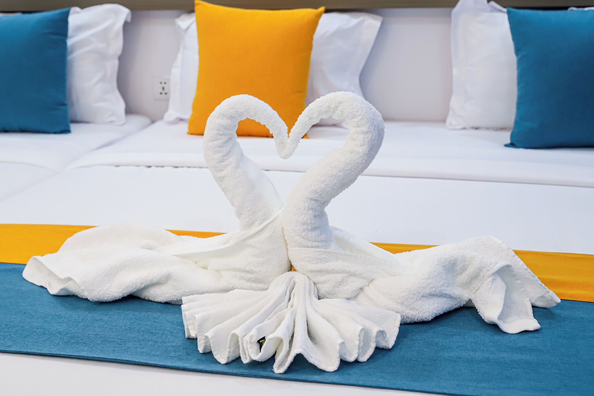 Un lit avec une literie blanche, un coussin bleu en guise d’élément décoratif, et une serviette pliée en forme de cœur.