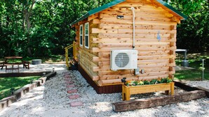 A wooden cabin with a green roof, a small porch, and a garden area with flowers. - #4 - Amish Log Camping Cabin Walk To The Water Cabin (Toronto)