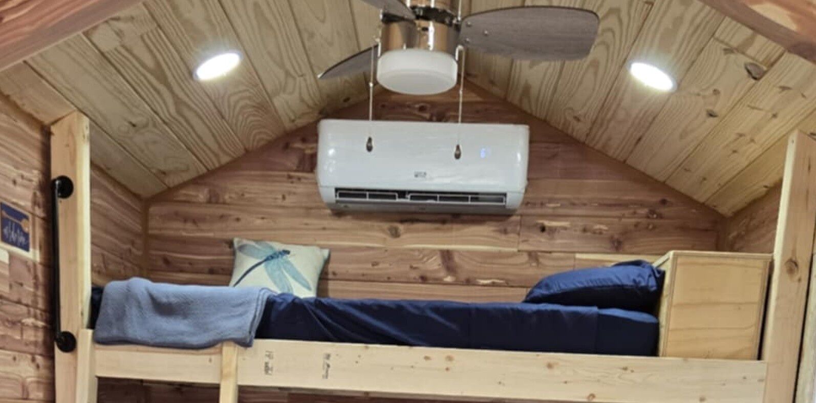 A small, wooden cabin-style room with a bunk bed, a ceiling fan, and an air conditioning unit.