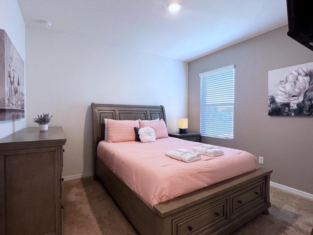 A bedroom with a bed, a nightstand, a dresser, a window with blinds, and a wall with a floral painting.