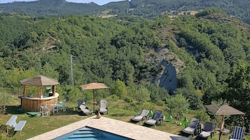 Zona de piscina con sillones de descanso y un bar, rodeada de vegetación exuberante y colinas.