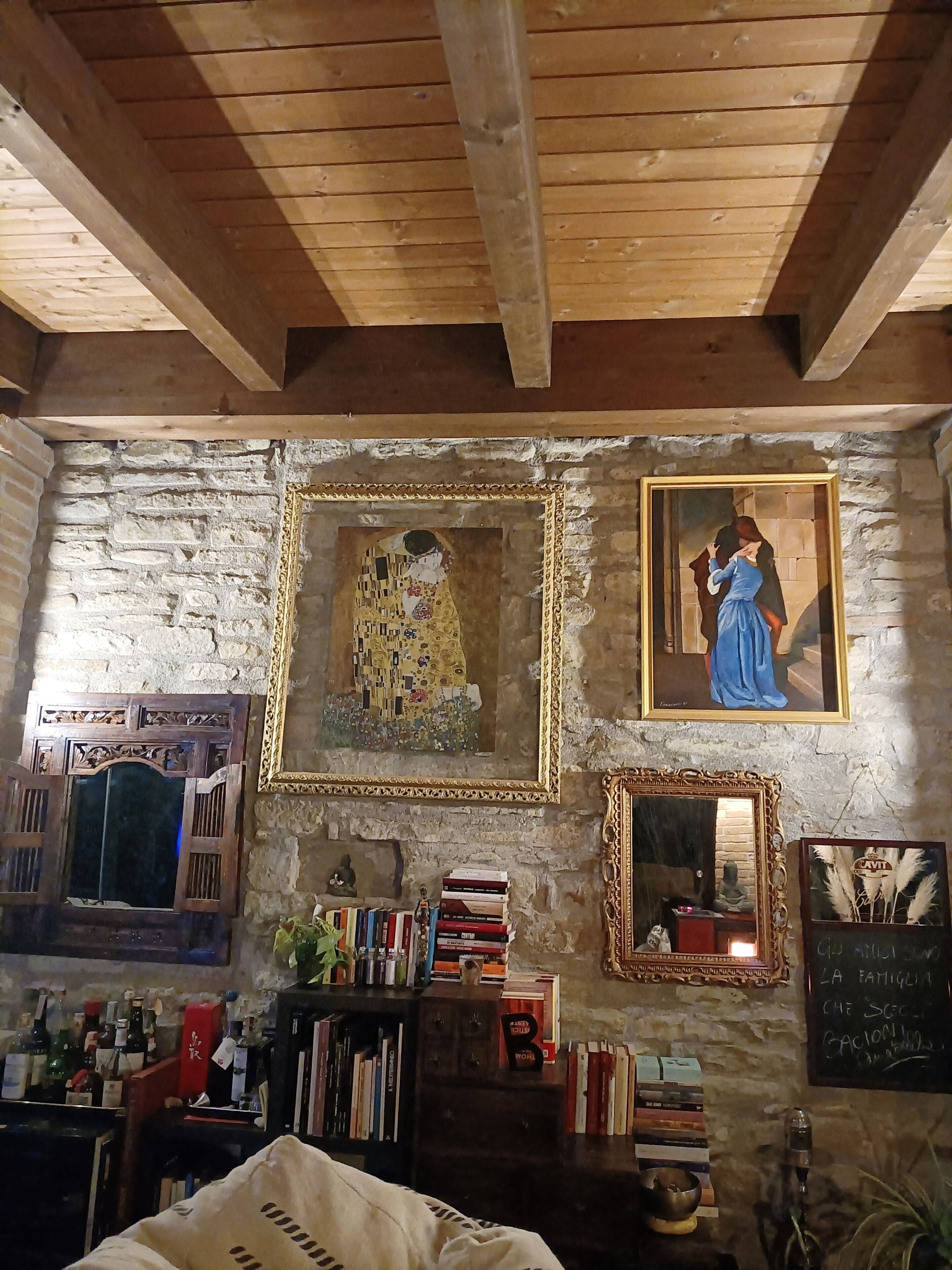 A room with exposed wooden beams, a stone wall, and two framed paintings. There is a bookshelf filled with books and decorative items.