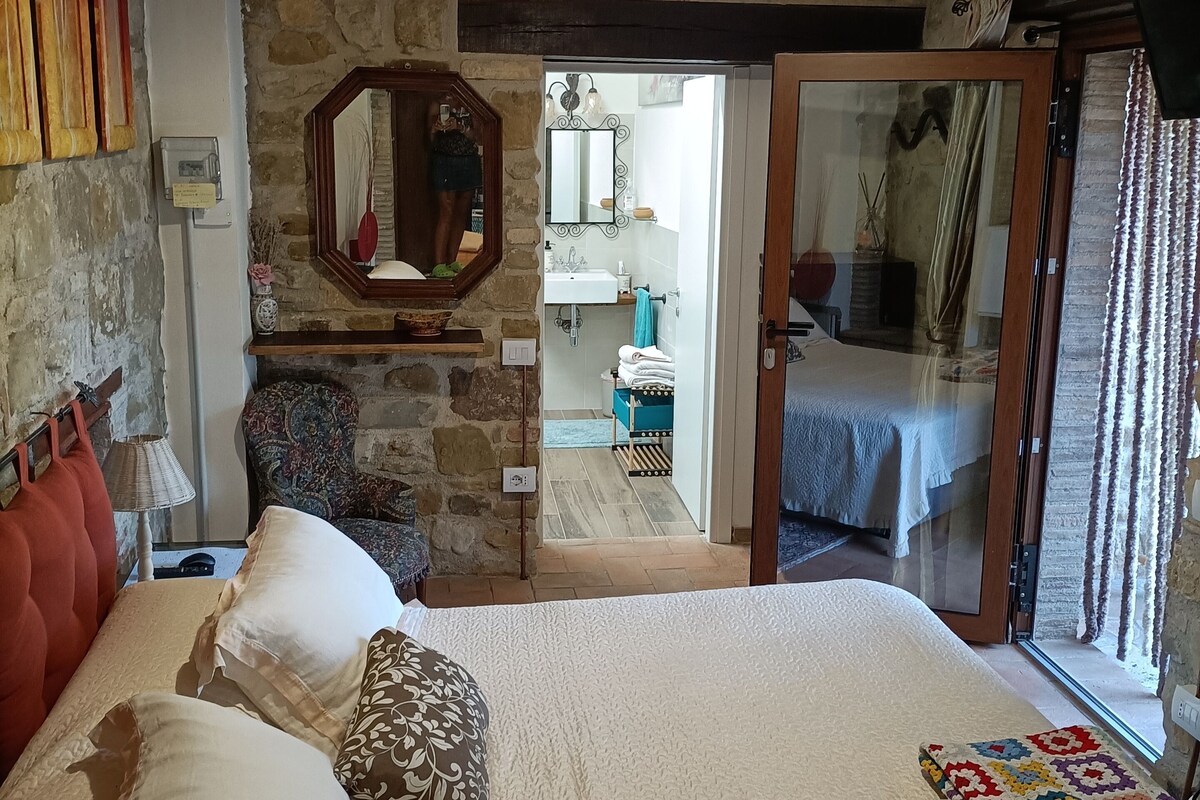 A bedroom with a stone wall, a bed with multiple pillows, a wooden headboard, a hanging light fixture, and a view into an adjacent room through a glass door.