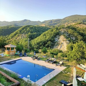 A pool area with lounge chairs and a wooden structure, surrounded by lush greenery and hills. - Casale I Calanchi Gubbio  (Gubbio)