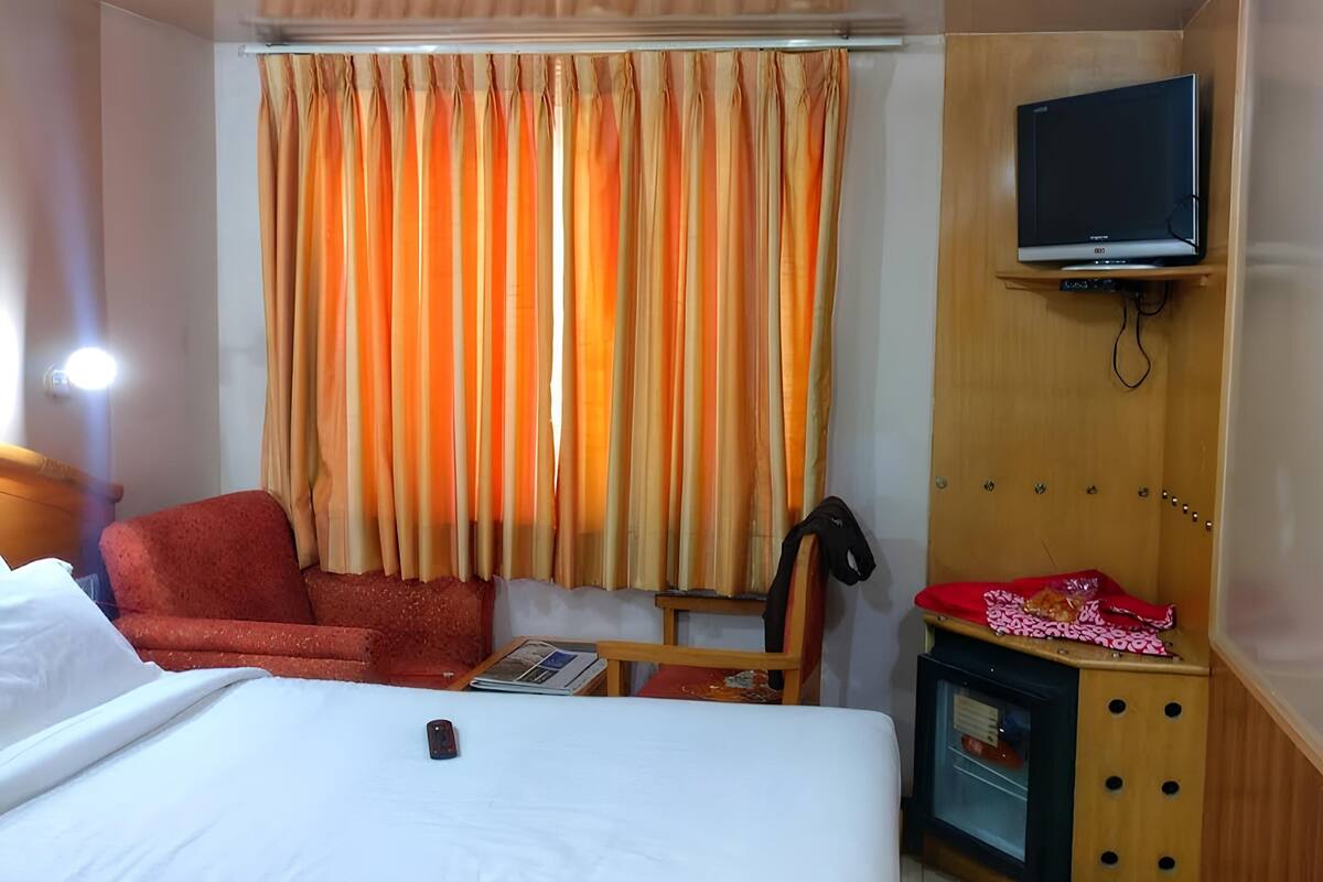 A hotel room with a bed, a red armchair, a television on a wooden stand, and a small table with a red cloth and snacks.