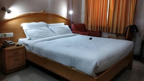 Un lit bien fait, avec du linge de lit blanc, une tête de lit en bois, une table de chevet avec un téléphone et une lampe, et une chaise rouge dans une chambre d’hôtel.
