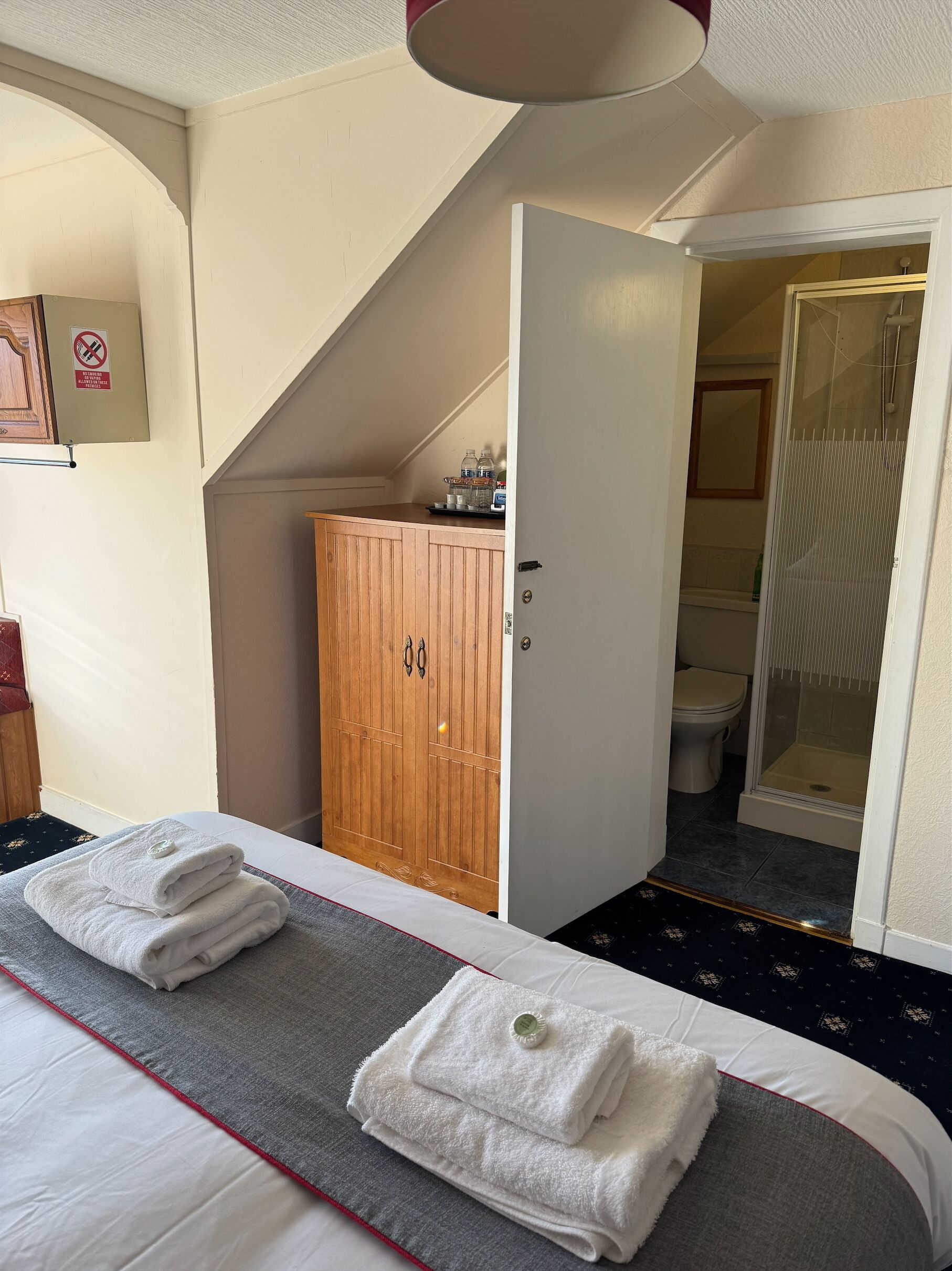 A hotel room with a bed, a wooden wardrobe, a bathroom with a shower, and a no-smoking sign on the wall.