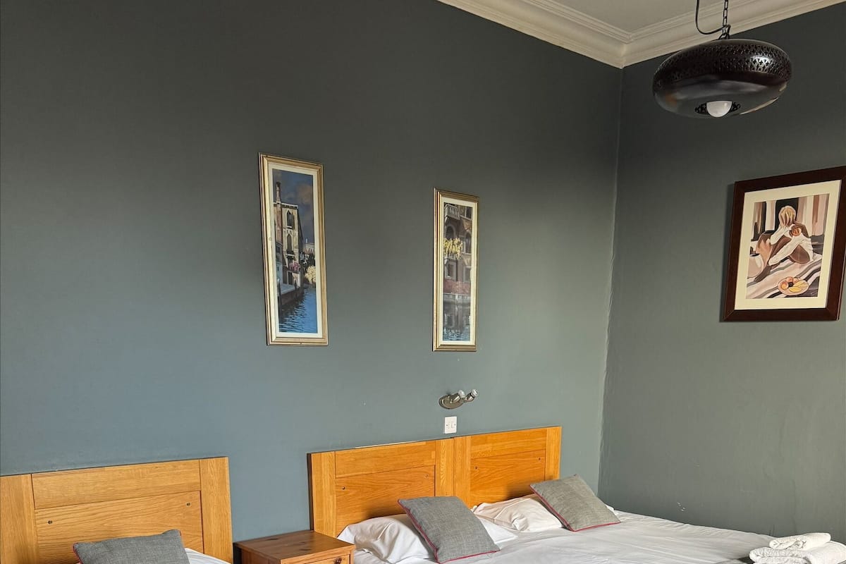 A hotel room with two beds, each with white bedding and grey blankets. There are framed pictures on the wall and a ceiling light fixture.