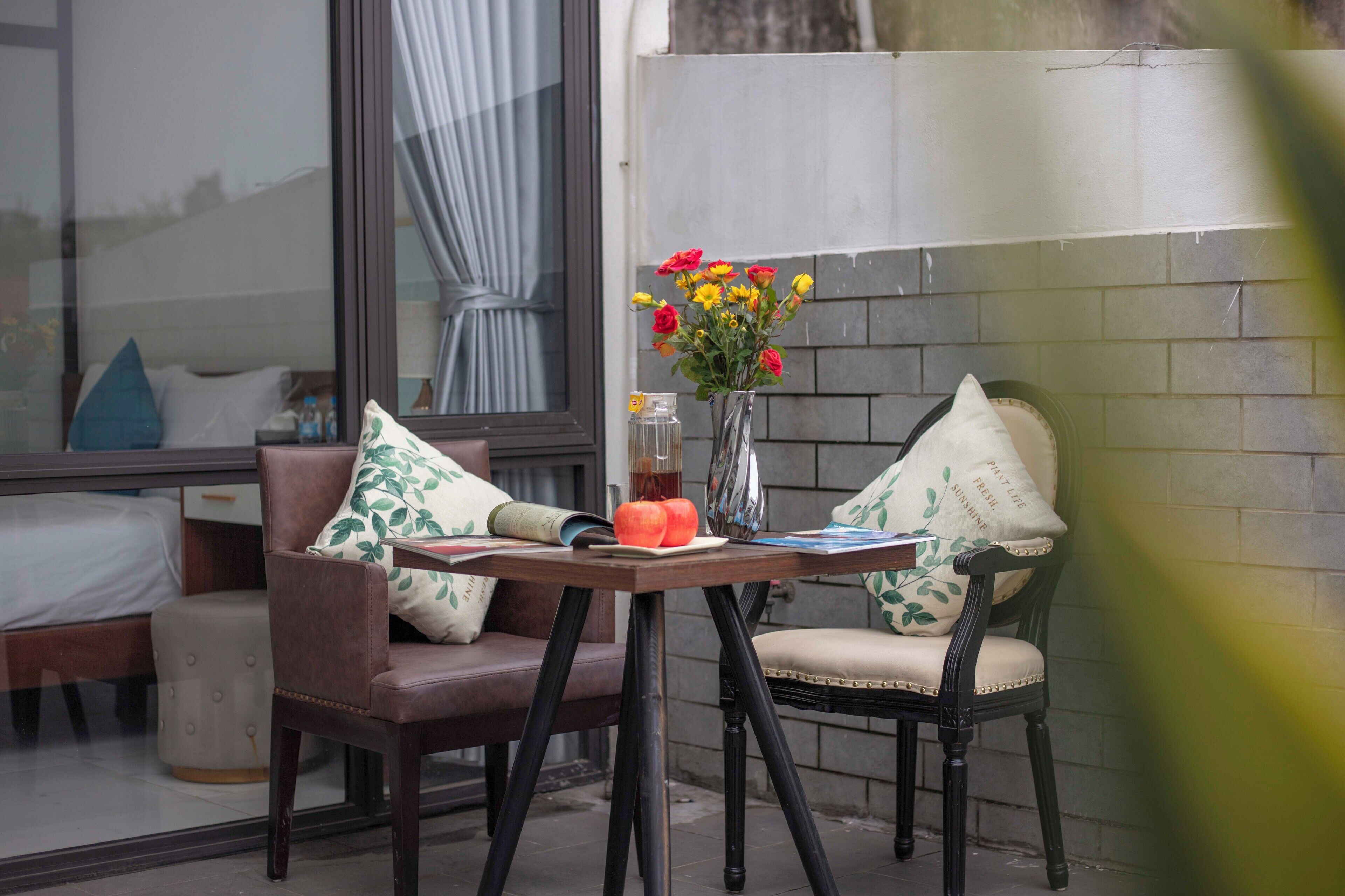 Un pequeño espacio al aire libre con una mesa y dos sillas, un florero con flores y una ventana con cortinas.