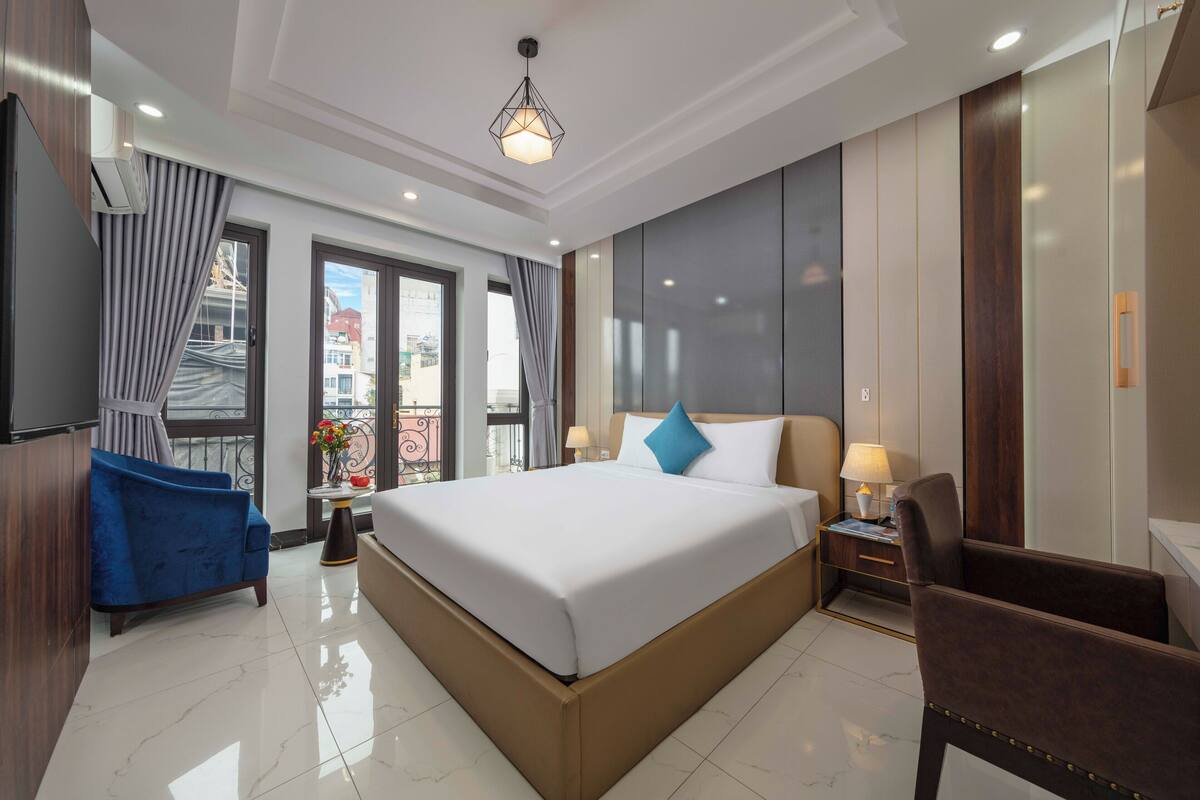 A hotel room with a large bed, a blue armchair, a small table with a lamp, and a view of buildings through the window.