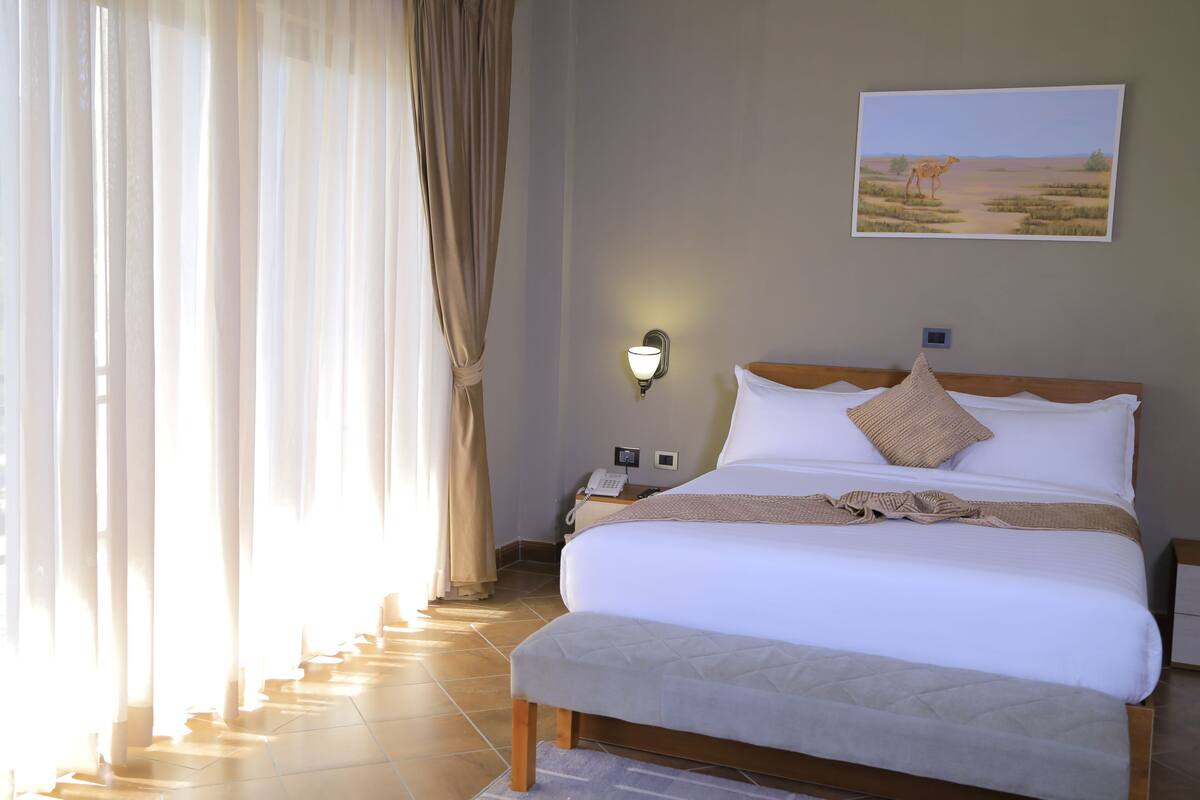 A hotel room with a large bed, a bedside table with a lamp, a painting of a desert scene, and a window with curtains.