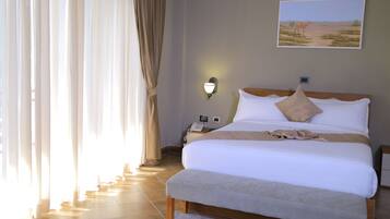 Ein Hotelzimmer mit einem großen Bett, einem Nachttisch mit Lampe, einem Gemälde, das eine Wüstenlandschaft zeigt, und einem Fenster mit Vorhängen.