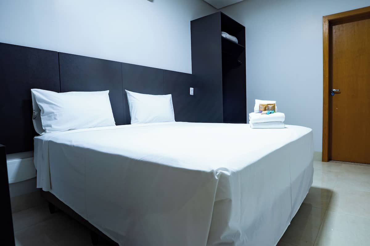 Un lit bien fait dans une chambre d’hôtel, avec une tête de lit sombre, une porte en bois et une petite table sur laquelle se trouvent un livre et une serviette.
