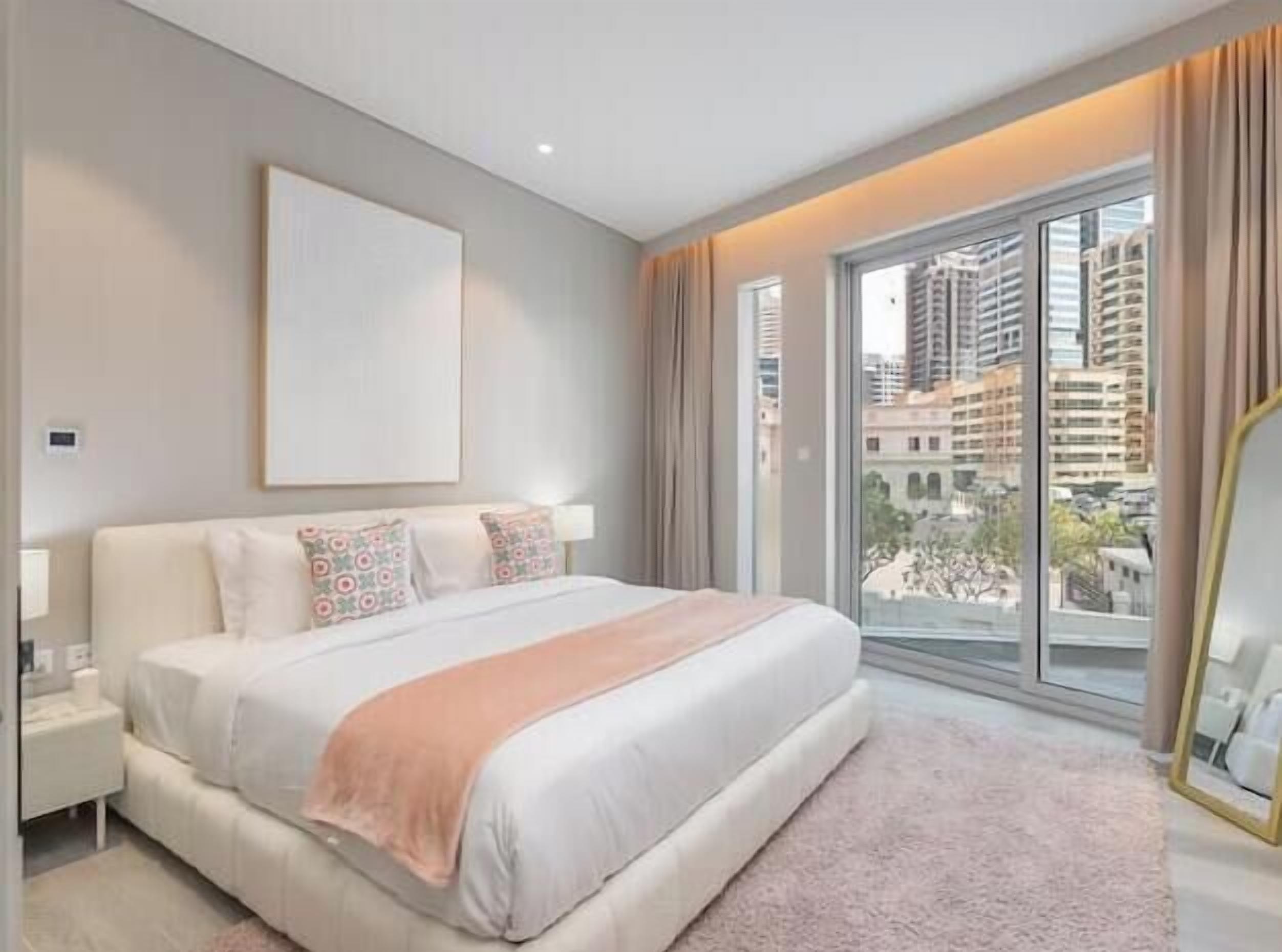 Une chambre d’hôtel moderne avec un grand lit, une table de chevet, une fenêtre donnant sur des immeubles et un miroir encadré.