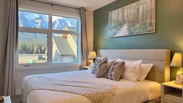 A bedroom with a large bed, a painting of a forest, and a window with a mountain view.