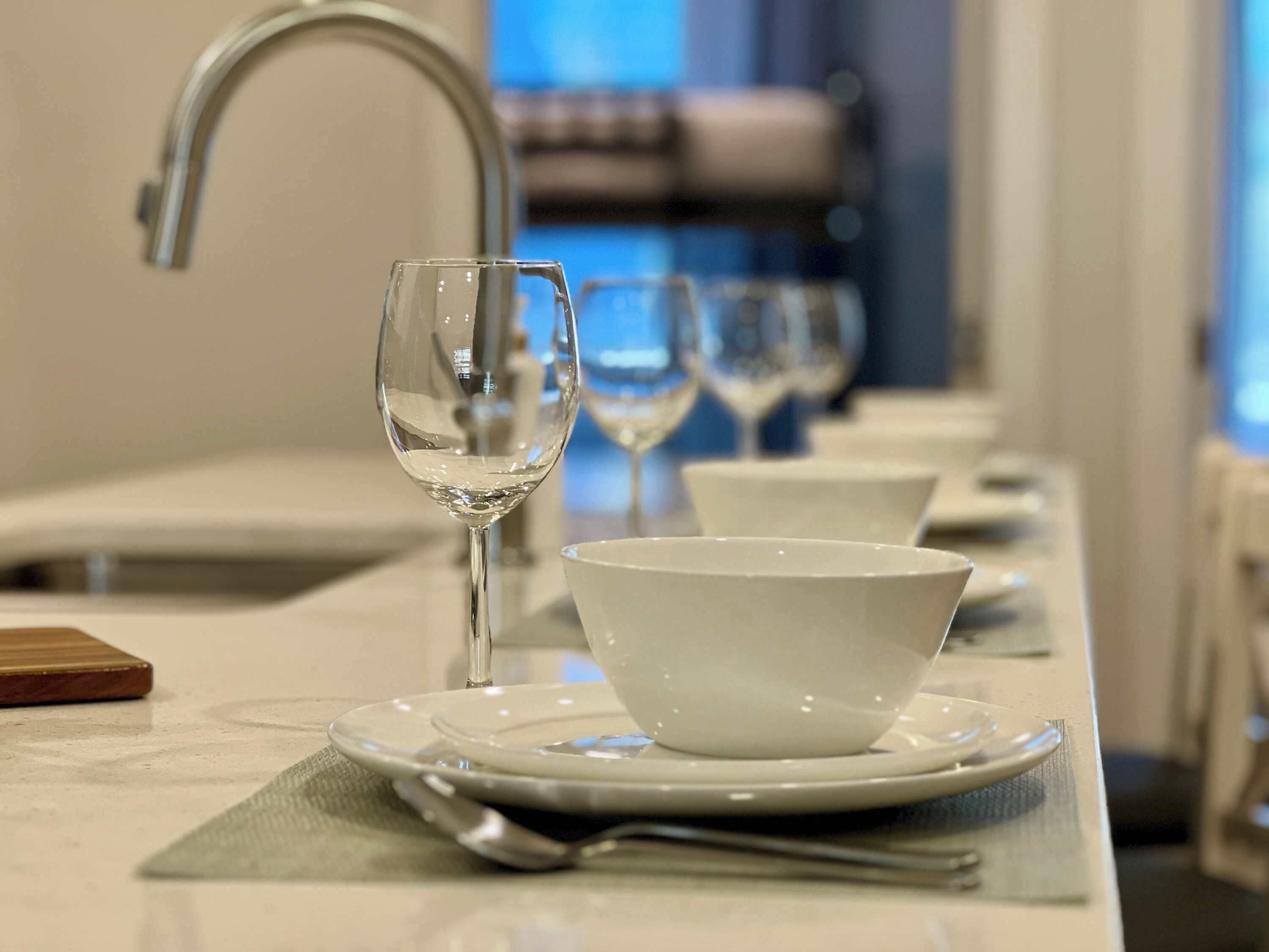 Un comedor ordenado con una mesa blanca, una copa de vino y un bol sobre un plato, con un lavamanos y un espejo al fondo.