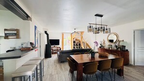 A spacious living area with a wooden dining table, comfortable seating, a staircase leading to an upper level, and a flat-screen TV mounted on the wall.