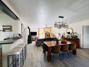 Uma sala de estar ampla com uma mesa de jantar de madeira, assentos confortáveis, uma escada que leva a um andar superior e uma TV de tela plana na parede.