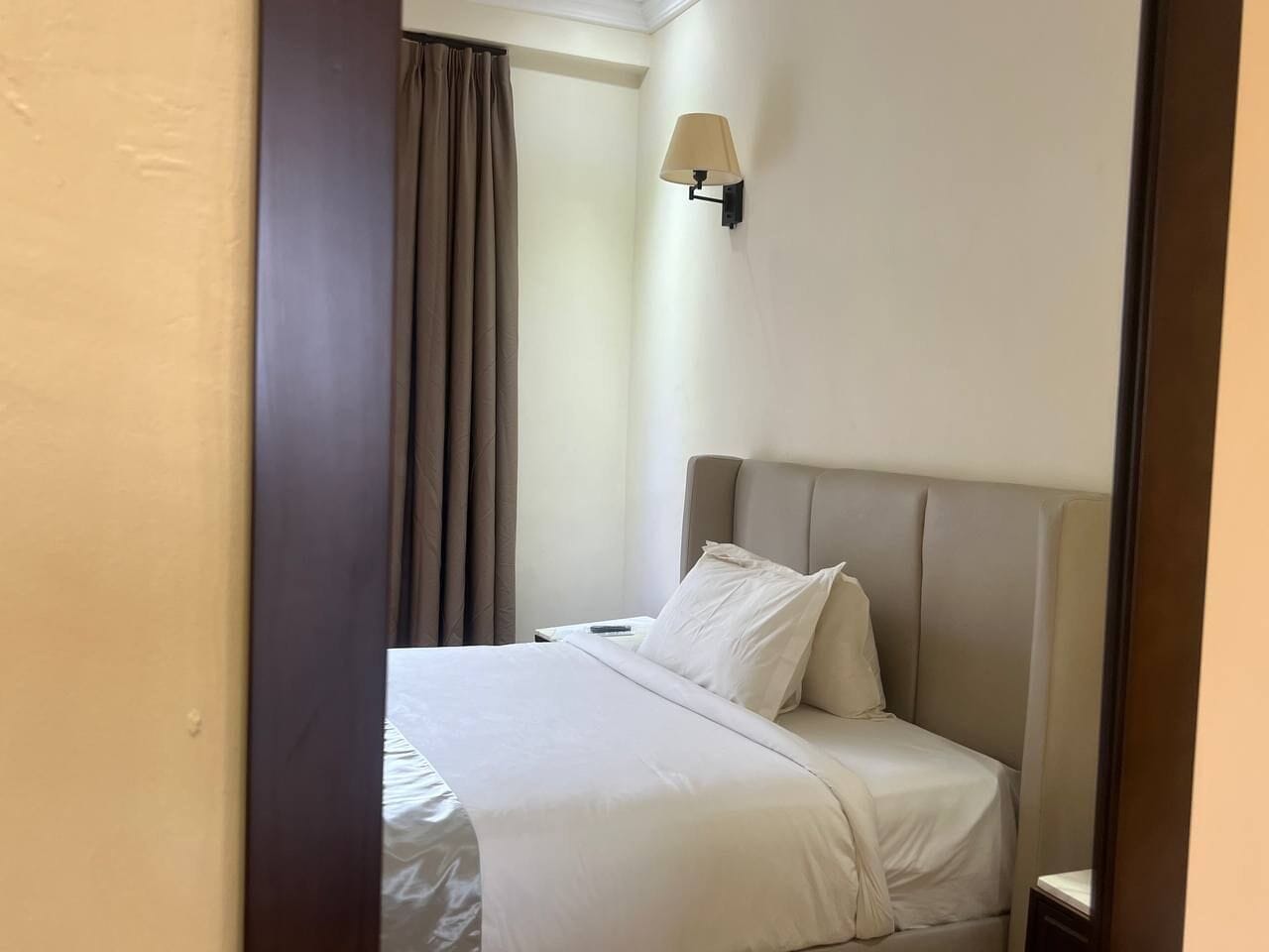 Una habitación de hotel con una cama, una mesita de noche, una lámpara y un ventanal con cortinas.