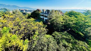 Un edificio moderno rodeado de exuberante follaje verde, con un cielo azul despejado y montañas a lo lejos.
