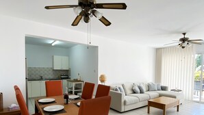A modern living room with a dining area, featuring a sofa, coffee table, and a dining table with chairs. There are ceiling fans and a kitchen visible in the background. - 70 PAP - Stay Anthea Gardens - Calm Living (Ayia Marinouda)