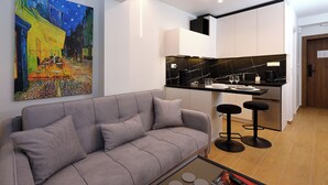 A modern living area with a grey sofa, a painting on the wall, a kitchen with black countertops, and two bar stools.