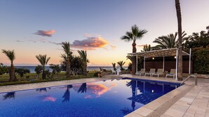 Piscina con vista al mar al atardecer, rodeada de palmeras y sillas de descanso.