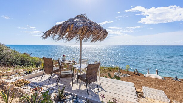 Una terraza de madera con sillas de mimbre y un toldo de paja con vista al mar.
