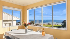 A dining area with a table set for two, large windows offering a view of the beach and ocean, and a vase with flowers on the table. - Gerroa House located mere steps from Seven Mile Beach. (Gerroa)