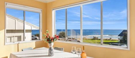 Un comedor con una mesa dispuesta para dos personas, ventanas amplias con vista a la playa y al océano, y un jarrón con flores sobre la mesa.