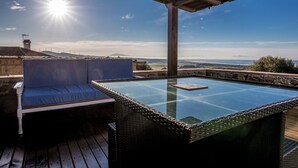 Un espacio al aire libre iluminado por el sol con una mesa de vidrio y un banco acolchado, con vistas a un paisaje pintoresco.