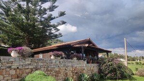 Una cabaña rústica de madera con un muro de piedra, rodeada de abundante follaje y plantas en flor.