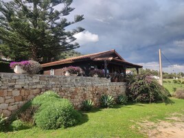 Una cabaña rústica de madera con un muro de piedra, rodeada de abundante follaje y plantas en flor.